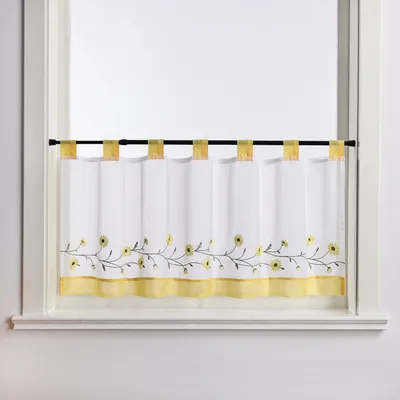 A close-up view of a white kitchen curtain with yellow floral embroidery and a yellow border, hanging on a black rod in a window frame.