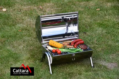 A portable charcoal grill filled with food, placed on a grassy lawn.
