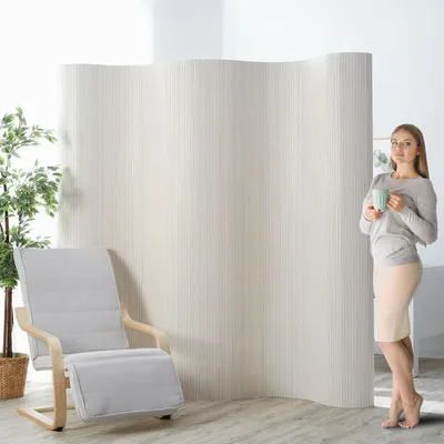 A woman stands holding a mug next to a room divider, with a chair and a plant visible.