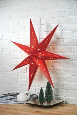 A red star-shaped decoration hangs on a white brick wall, with two small Christmas trees and a teapot on a tray below.