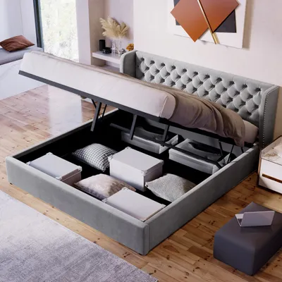 A grey upholstered bed with its storage compartment open, revealing pillows and storage boxes inside. The bed is in a room with wooden flooring and a window in the background.