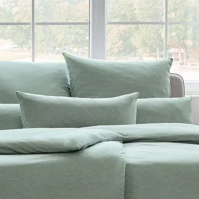 A close-up view of several mint green pillows and bedding arranged on a bed or sofa, with a blurred window and trees in the background.