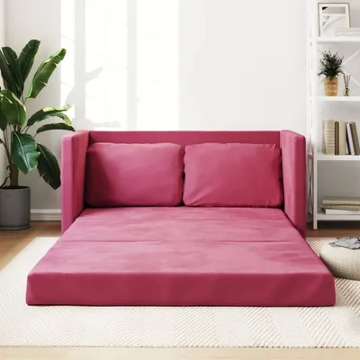 A pink sofa bed with two cushions is laid out on a light-colored rug in a well-lit room. A large potted plant is visible on the left, and a white shelf unit is in the background on the right.