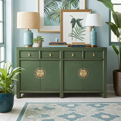 A green sideboard with decorative brass accents is placed in a room with light blue walls. It is decorated with two table lamps, books, and framed botanical prints. A potted plant is on the left, and a large potted plant is on the right. A patterned rug is