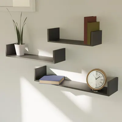 Three dark gray U-shaped shelves mounted on a light gray wall. The top shelf holds a potted plant and two books. The middle shelf holds two books. The bottom shelf holds a book and a clock.