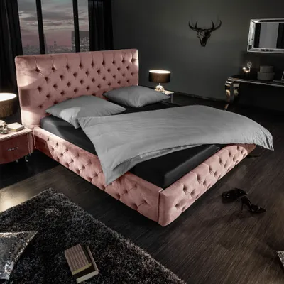 A luxurious bedroom scene featuring a pink velvet Chesterfield bed with grey bedding, a dark rug, and ambient lighting.