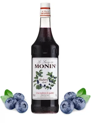 A bottle of Monin Blueberry syrup is shown upright, with blueberries and leaves arranged around its base.