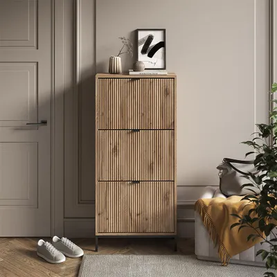 A narrow shoe cabinet with three flip-down drawers stands in a hallway next to a door and a bench with a blanket.