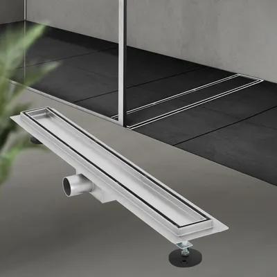 A silver linear shower drain with black rubber seals is shown in the foreground, angled diagonally. The background shows a modern bathroom with dark gray tiled flooring and a glass shower enclosure.