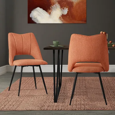Two orange dining chairs are placed around a small dark wood table with a green cup on it. A painting hangs on the wall behind them.