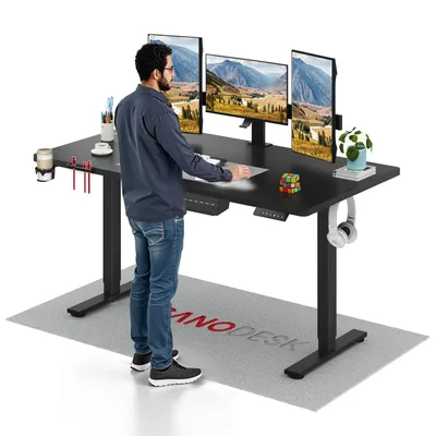 A man stands at a black standing desk with three monitors, a cup holder, and headphones. A mat with a logo is on the floor.