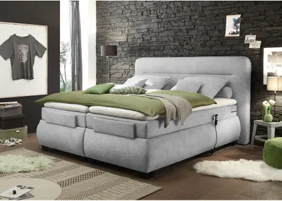 A bedroom scene featuring a large, adjustable box spring bed with green bedding and pillows, set against a dark stone wall.