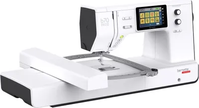 A white and gray bernette b70 DECO sewing machine with an embroidery module attached, shown from a diagonal front-left angle against a white background.