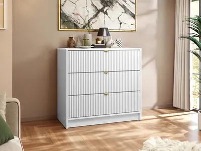 A white dresser with gold handles is shown in a room with a painting on the wall and a plant in the background.