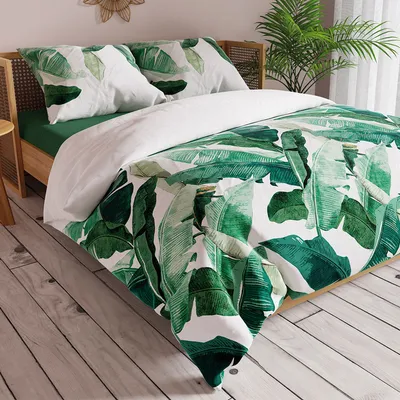 A bed with green and white banana leaf patterned bedding, set in a bedroom with wooden flooring and a plant in the background.
