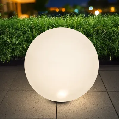 A glowing spherical lamp sits on a tiled patio in front of a dense green hedge at dusk.
