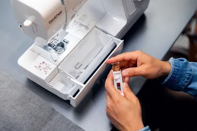 A person's hands are shown holding a bobbin and bobbin case near an open sewing machine.