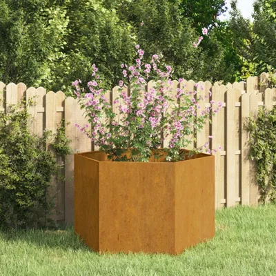 A hexagonal planter made of Corten steel with a rusty finish, containing a flowering plant with pink blossoms, set in a garden with a wooden fence in the background.