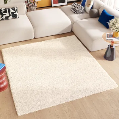 A cream-colored rug is placed on a wooden floor in front of a modern, modular white sofa. Decorative pillows in black and white, blue, and beige are on the sofa. A small side table with a vase of flowers and a book is next to the sofa.