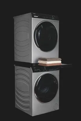A stacked washing machine and dryer unit with a shelf and folded towels between them, set against a black background.