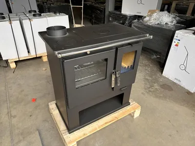 A black wood-burning stove with an oven and cooktop is displayed on a wooden pallet in what appears to be a warehouse or storage area. Other products and packaging are visible in the background.
