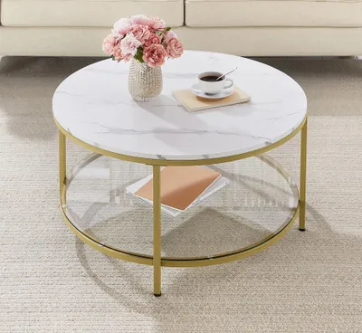 A round coffee table with a marble-like top and gold legs, placed on a rug in a living room setting. A vase of pink flowers and a cup of coffee are on the table.