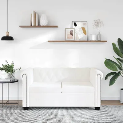 A white Chesterfield sofa is shown in a well-lit room with decorative shelves and a plant.