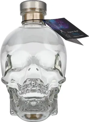 A clear glass skull-shaped bottle filled with clear liquid, with a silver cap and a tag attached to the neck.
