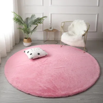 A round pink rug with a black and white polka dot pillow on it, in a room with a plant and a chair.
