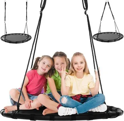 Three young girls are sitting on a black round swing, smiling at the camera. The swing is suspended by ropes from above, and two other identical swings are visible in the background.