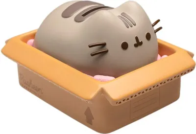 A Pusheen the cat figurine is sitting in a cardboard box filled with pink packing peanuts.