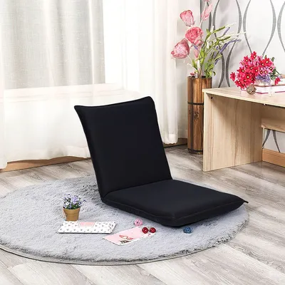 A black floor chair is placed on a round gray rug in front of a window and a desk with flowers.