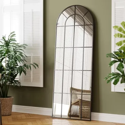 A tall, arched mirror with a black metal frame stands in a room with green walls and white shutters. A potted plant is to the left, and a wicker chair is reflected in the mirror.
