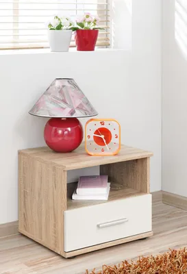 A bedside table with a lamp and a clock on top, and books in the open shelf.