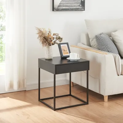 A black side table with a drawer is placed next to a beige sofa in a living room. The table has a vase with dried flowers, a framed picture, and a cup of coffee on it. A window with white curtains is visible in the background.