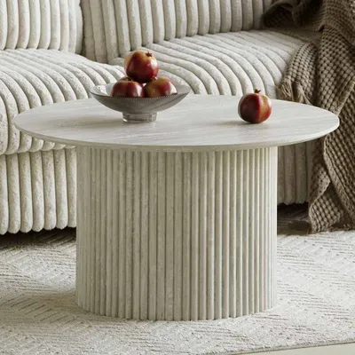 A round coffee table with a fluted base and a marble-like top, decorated with a bowl of apples and a single apple, placed on a rug in front of a sofa.