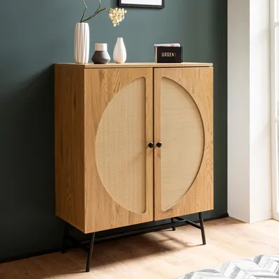 A wooden sideboard with rattan woven doors stands against a dark green wall, with decorative items on top.