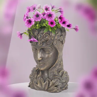 A bronze-colored planter shaped like a woman's head, filled with purple petunias, sits on a white surface against a blurred pink and white background.