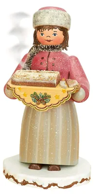 Ein fröhliches Mädchen in traditioneller Kleidung hält ein Tablett mit einem Stollen in den Händen während Rauch aus ihrem Kopf aufsteigt was darauf hindeutet dass es sich um ein Räuchermännchen handelt das auf einem schneebedeckten Sockel steht