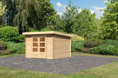 A wooden garden shed with a green roof sits on a paved area surrounded by lush greenery and flowers.