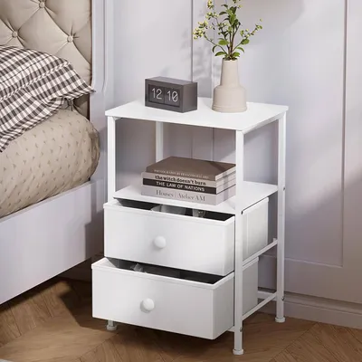 A white bedside table with two drawers, a clock, books, and a vase with flowers.