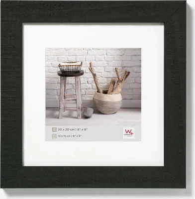 A framed picture with a white mat, showing a rustic stool with a basket on top, a decorative pot with branches, and a brick wall in the background.