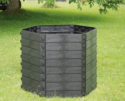 A black hexagonal compost bin sits on a grassy lawn with a tree and foliage in the background.