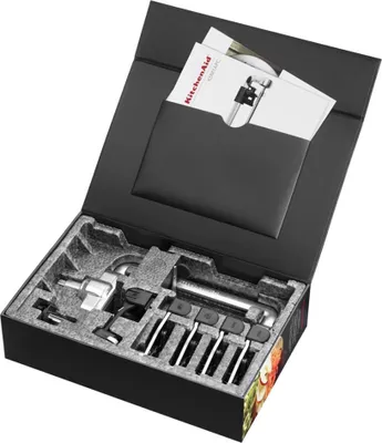 A black box opened to reveal KitchenAid food processor attachments nestled in molded foam packaging. Manuals are tucked into a black pocket on the inside lid.