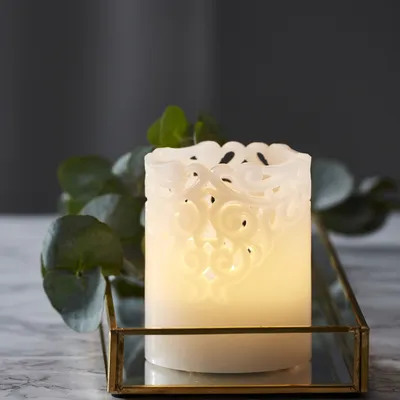 A white LED candle with a decorative filigree design sits on a gold-trimmed glass tray, next to some green foliage.