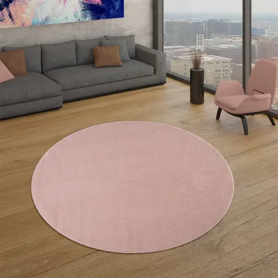 A modern living room with a gray sectional sofa, a pink armchair, and a large abstract painting on the wall. A round pink rug is placed on the wooden floor in the foreground.
