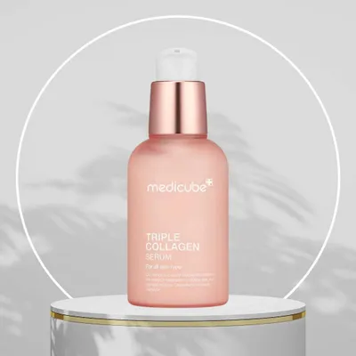 A bottle of Medicube Triple Collagen Serum is displayed on a white pedestal against a light gray background with circular white framing and subtle shadow effects.