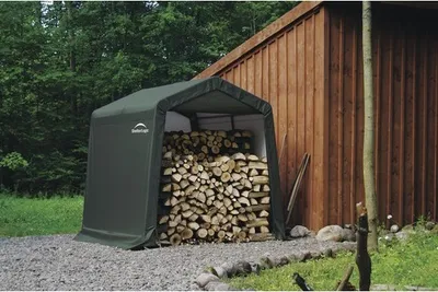 A dark green storage tent filled with firewood is parked next to a wooden building in a wooded area.