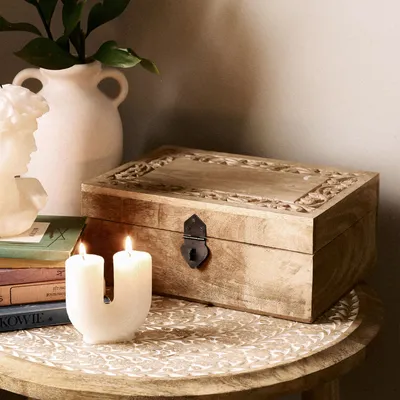 A wooden box with carved details sits on a round table next to two white candles and a stack of books. A white vase with greenery is in the background.