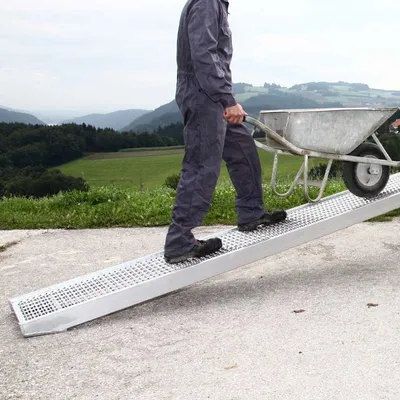 A person in a blue jumpsuit pushes a wheelbarrow up a ramp in a rural setting.